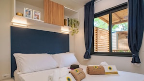Hotelkamer met een bed, witte kussens, een hoofdbord, een raam met gordijnen, een plank met ingekaderde foto's en handdoeken met een logo.