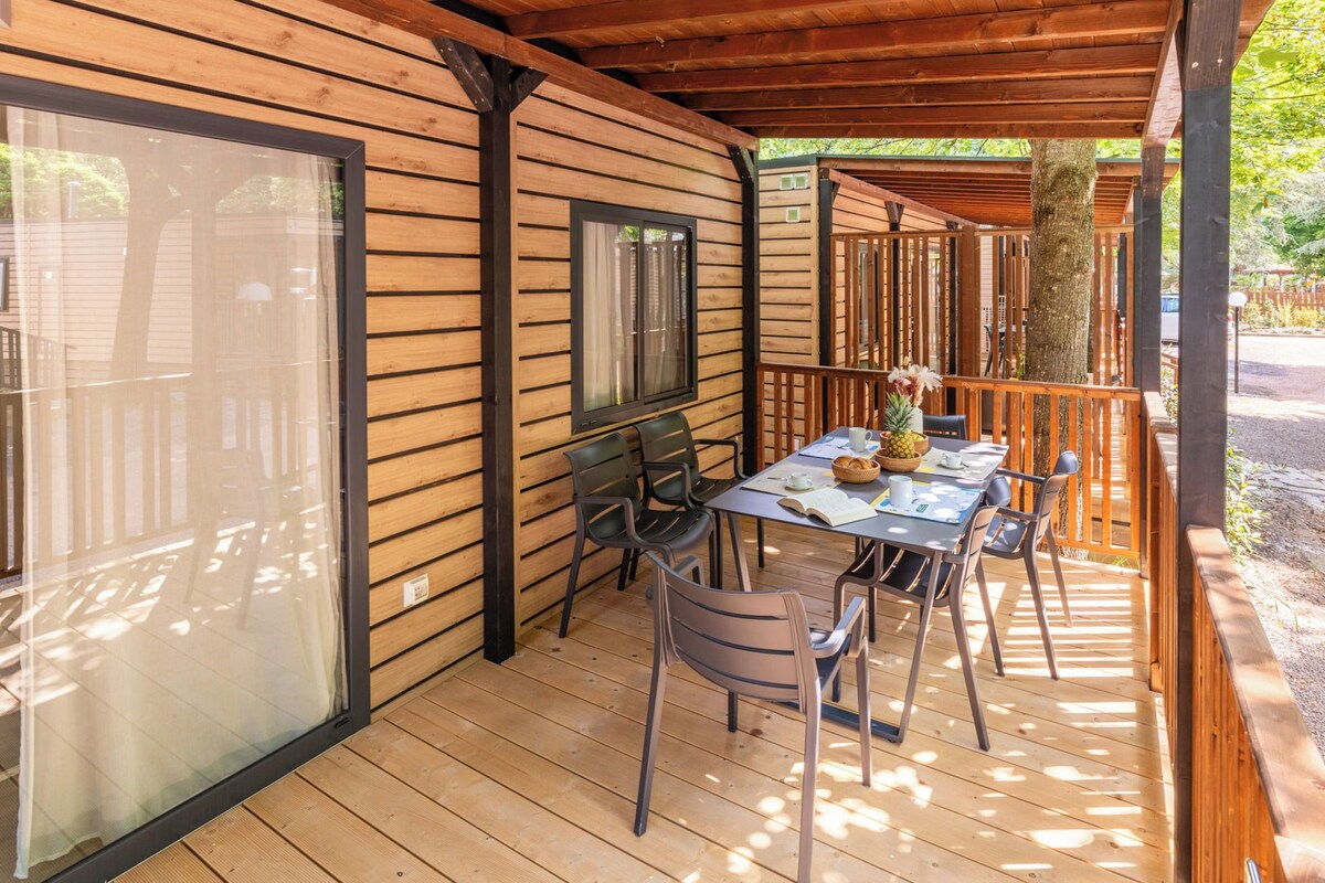 Un espace extérieur abrité, doté d’une terrasse en bois, d’une table avec des chaises et offrant une vue sur les arbres environnants.