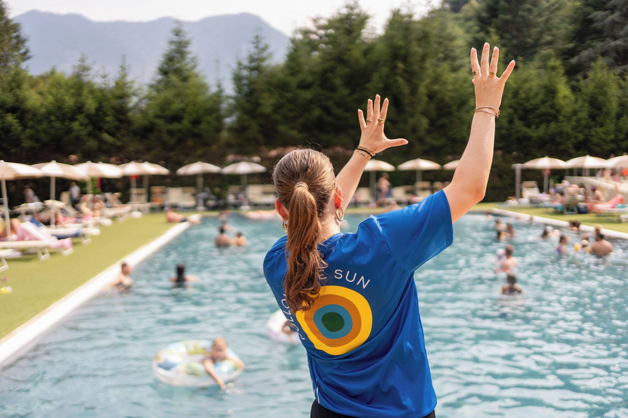 Une personne portant une chemise bleue avec le mot « SUN » écrit dans le dos, se tient près d’une piscine où des gens nagent et prennent des bains de soleil.