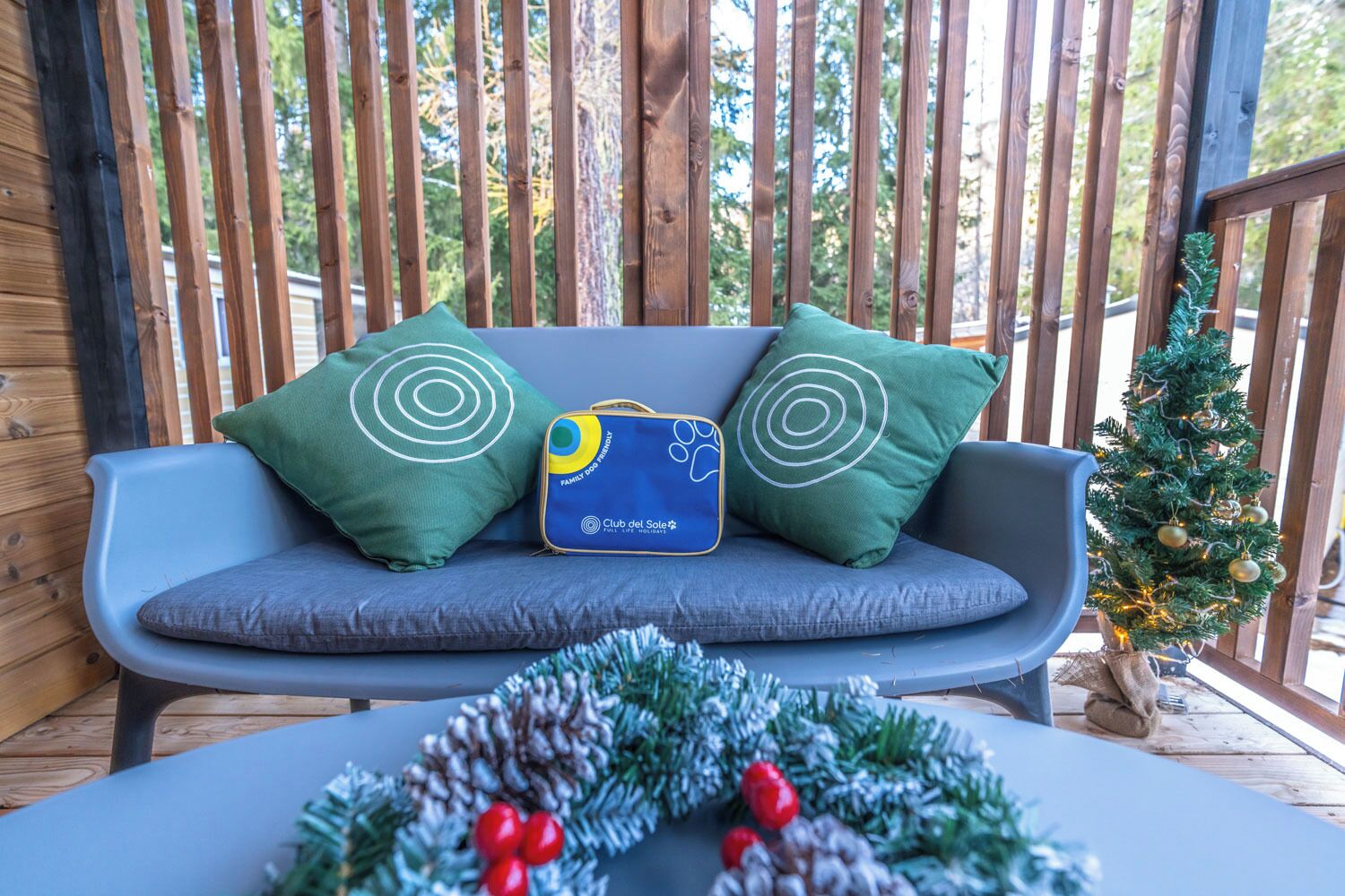 A wooden deck with a blue sofa, green pillows, a small Christmas tree, and a decorative wreath.