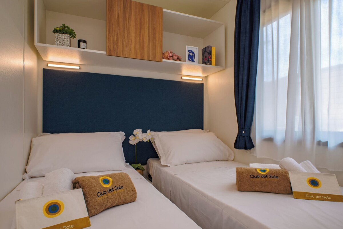 Une chambre d’hôtel avec deux lits, chacun agrémenté d’un oreiller portant l’inscription « Club del Sole ». On y trouve des tables de chevet avec des lampes, une étagère au-dessus des lits et une fenêtre avec des rideaux.