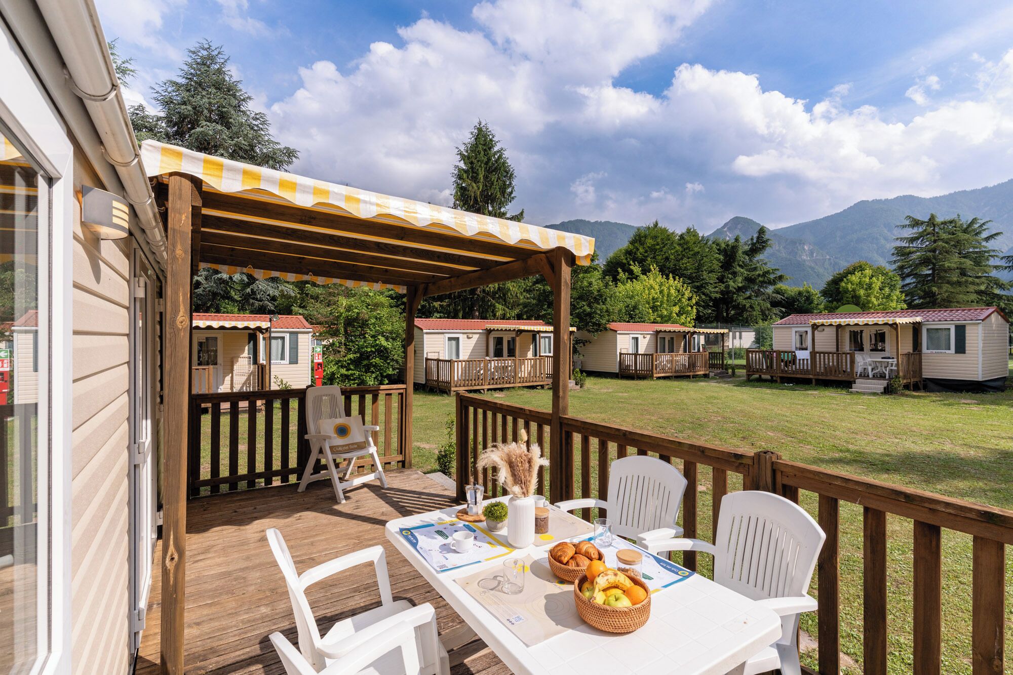 A wooden deck with a table set for breakfast, surrounded by white chairs, overlooking a grassy area with mobile homes and mountains in the background.