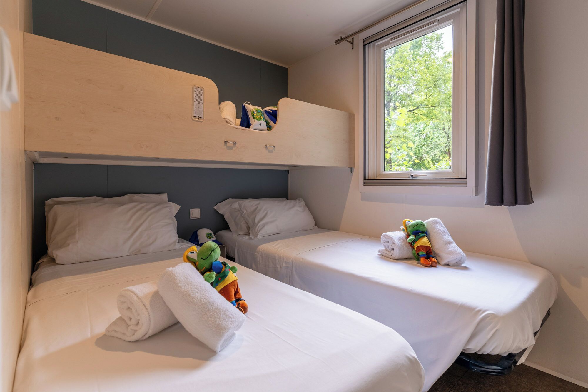 Een kamer met twee bedden, een raam met gordijnen en een inbouwplank boven de bedden.