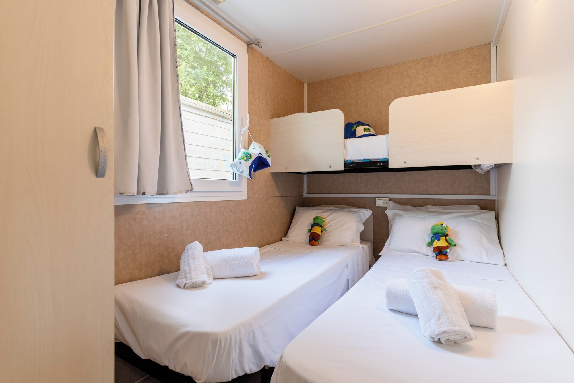 A small, single-room accommodation with two beds, a window with curtains, and a bunk bed above.