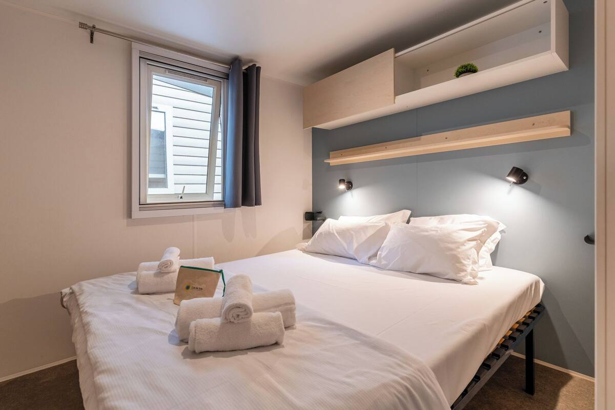 Een hotelkamer met een bed, handdoeken, een nachtkastje, een raam met gordijnen en inbouwkasten.