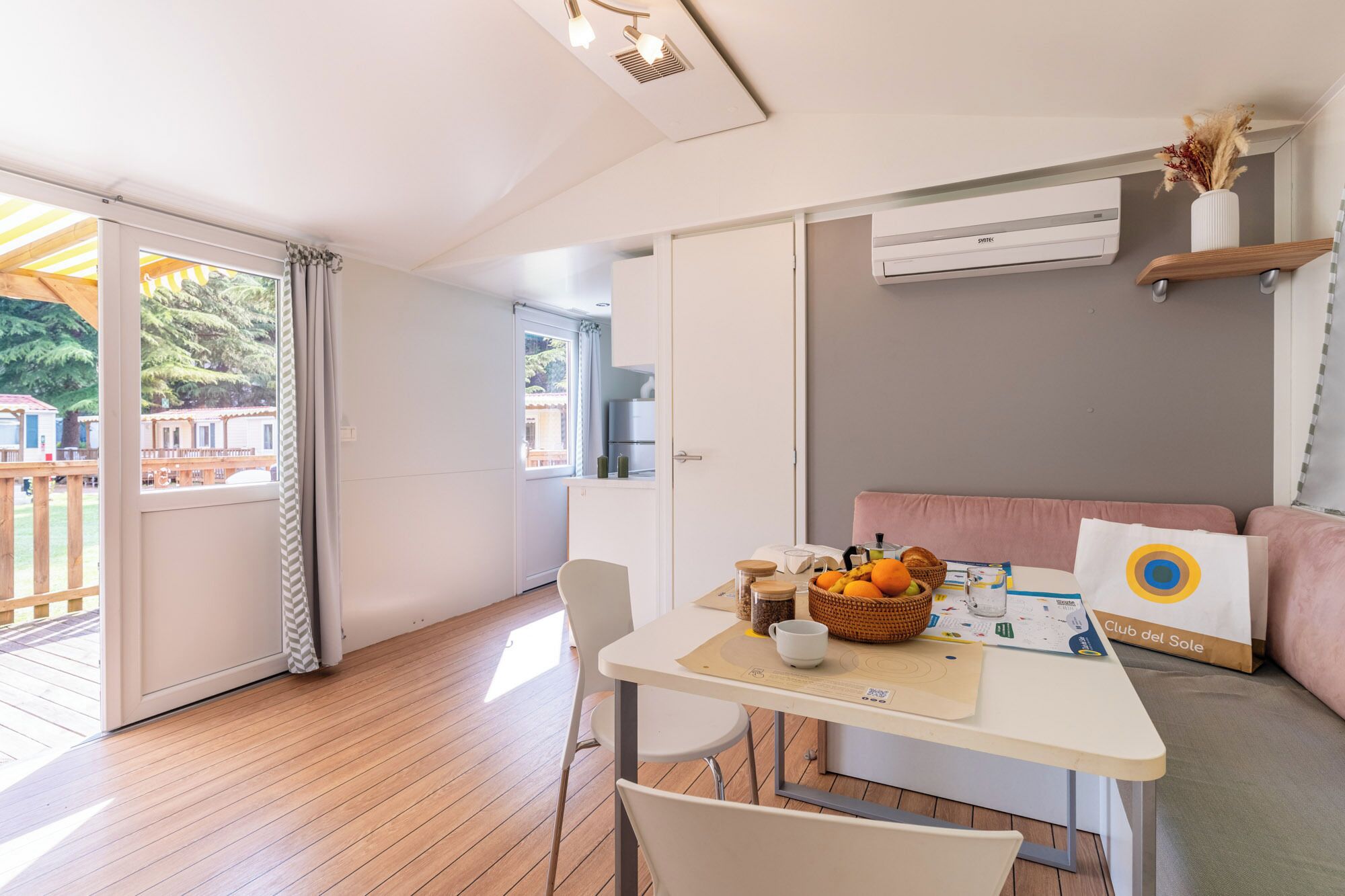 Une pièce avec un plancher en bois, une table sur laquelle se trouve un panier de fruits, une chaise blanche, un canapé avec un coussin et un appareil de climatisation.