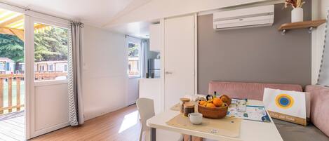 Una habitación con piso de madera, una mesa con una cesta de fruta, una silla blanca, un sofá con un cojín y un equipo de aire acondicionado.