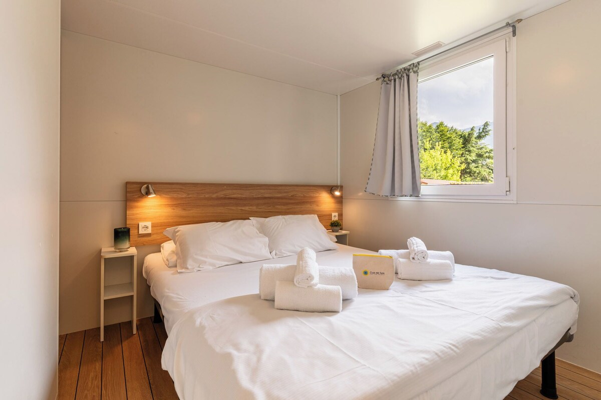 A hotel room with a wooden headboard, a bed with white linens, a nightstand with a lamp, and a window with a view of trees.