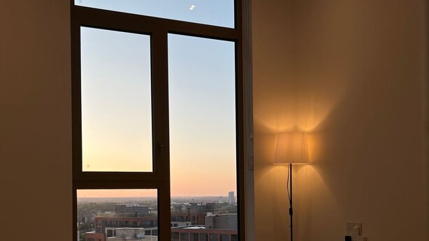 A bedroom with a large window, a bed, a floor lamp, and a view of a cityscape.