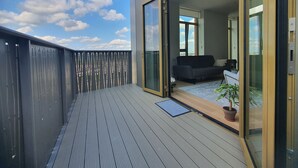 A balcony with a wooden floor, a sliding glass door leading to a living area with a sofa and a potted plant.
