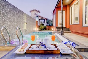 A breakfast tray with pastries, coffee, tea, and juice by a poolside with a stone wall and a building in the background. - Elivaas Irene Jade 3BHK Pvt Pool Garden with Balconies (Mawal)
