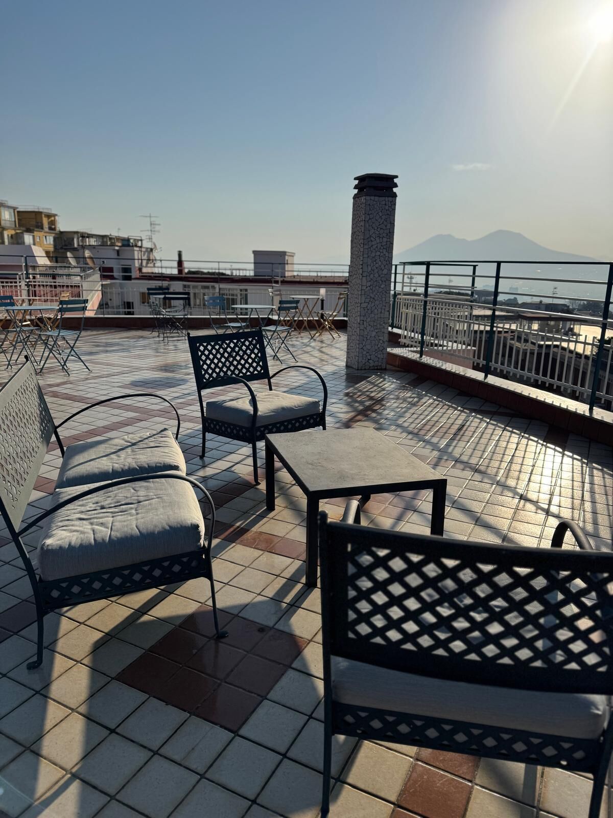 A rooftop terrace with outdoor seating, a stone pillar, and a view of buildings and mountains in the distance.