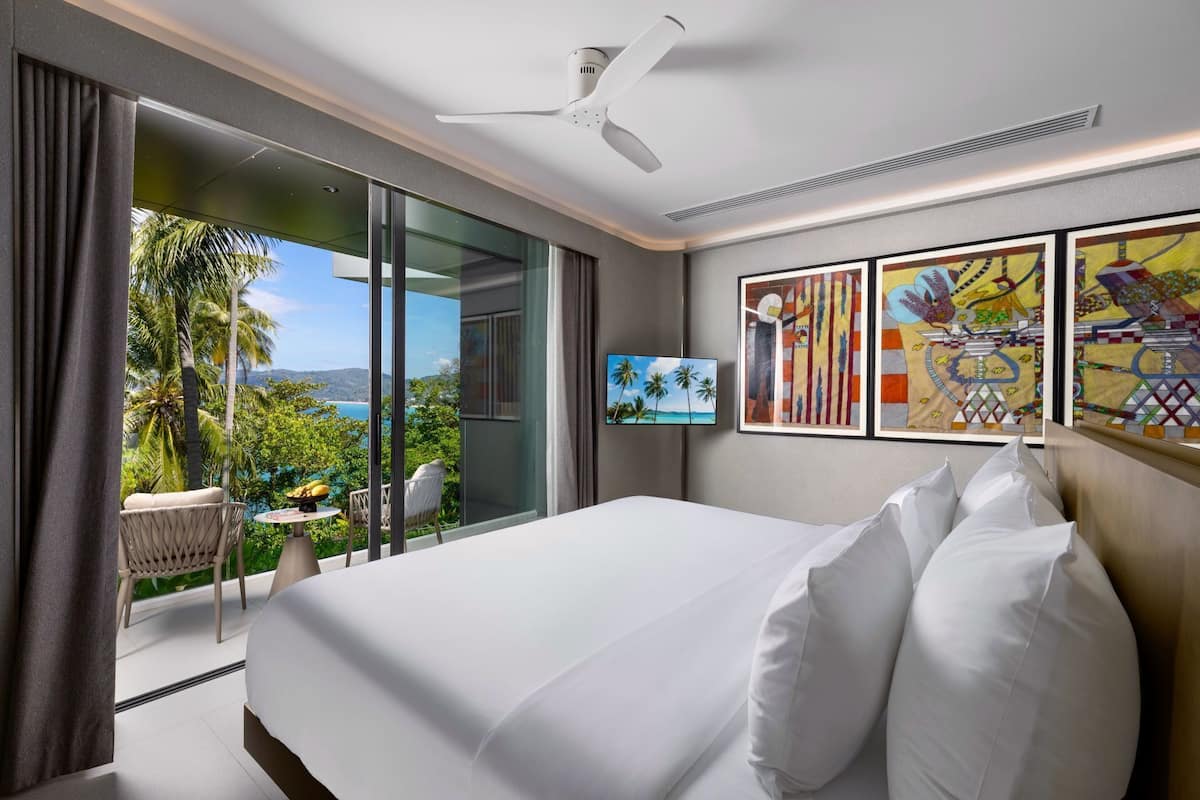 A hotel room with a large bed, a ceiling fan, a TV, and a balcony with a view of palm trees and the ocean.