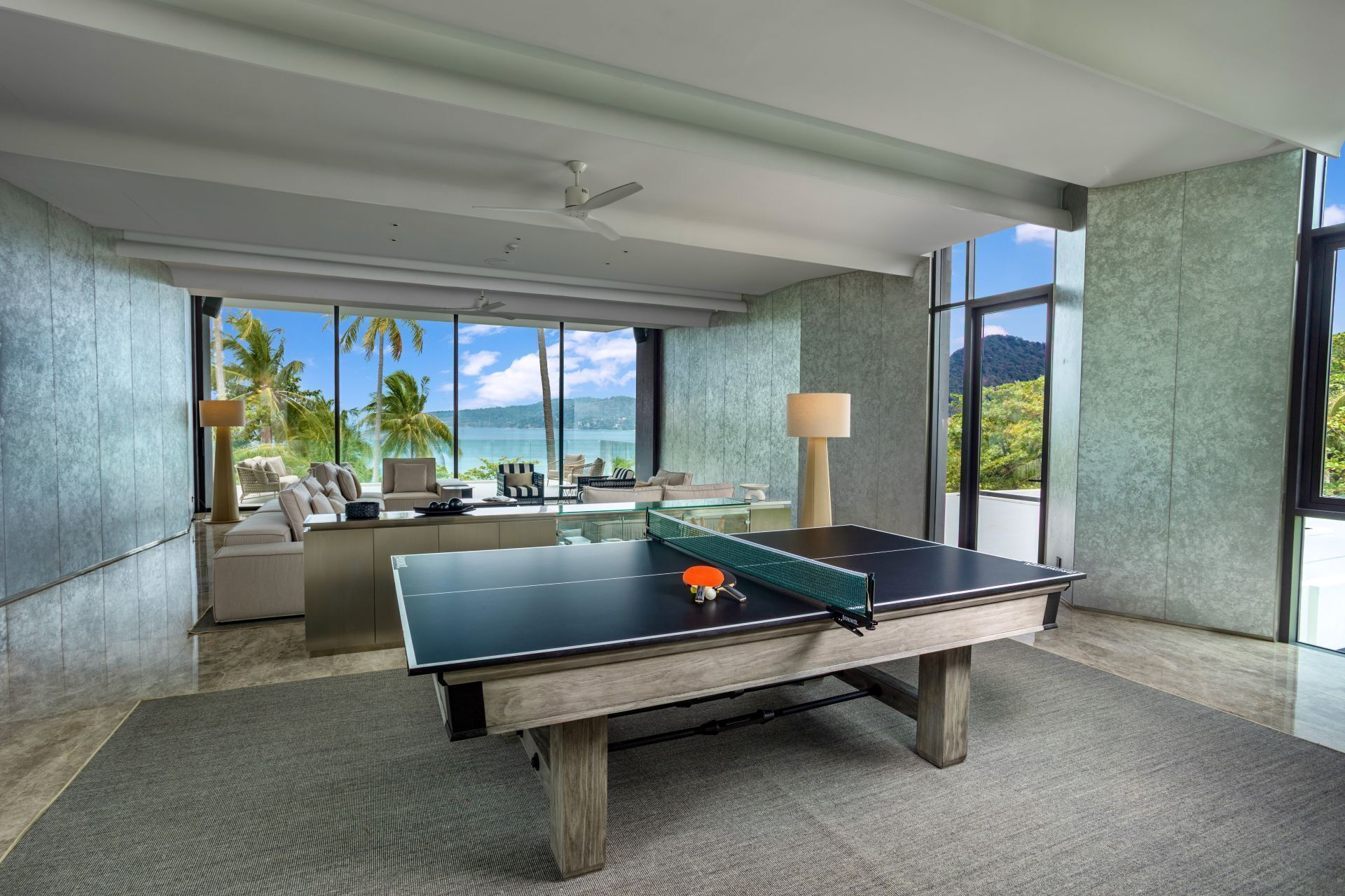 Una habitación amplia con mesa de ping pong, ventanas grandes que ofrecen vista a palmeras y al océano, y un diseño interior moderno.
