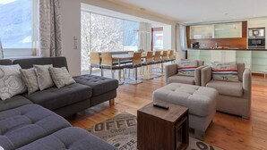 Een moderne woonkamer met een hoekbank, een klein voetenbankje en een salontafel. Er is een eetgedeelte met een houten tafel en stoelen, en een keuken met een magnetron en kasten.