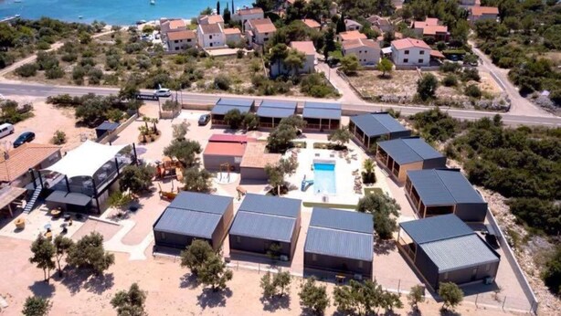 Vista aèria d'una estació turística a la costa, amb diversos edificis, una piscina i una vista clara del mar.