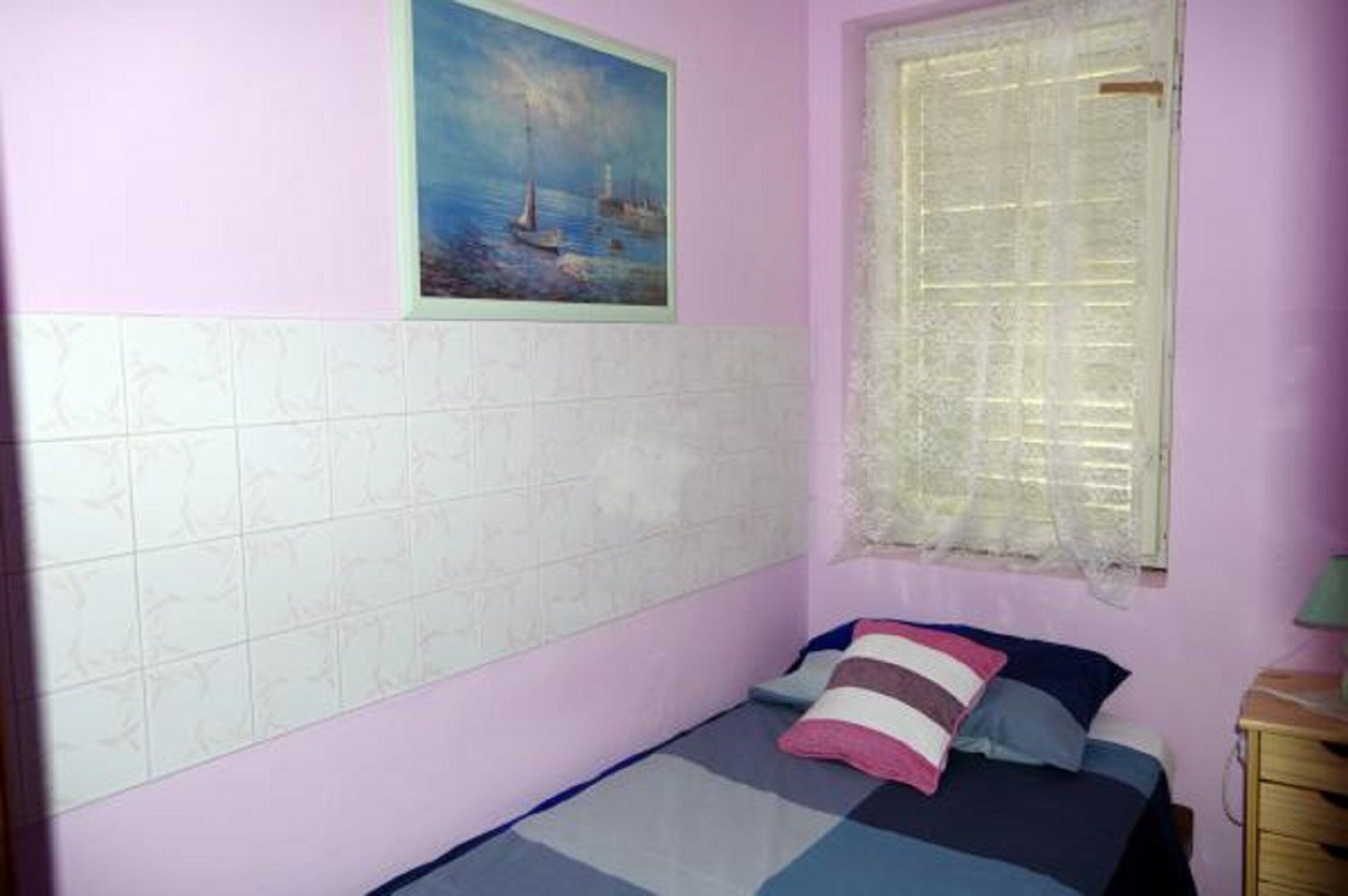 Ein Schlafzimmer mit einem Bett, einem Fenster mit Spitzenvorhängen, einem Meeresbild und einer gefliesten Wand.