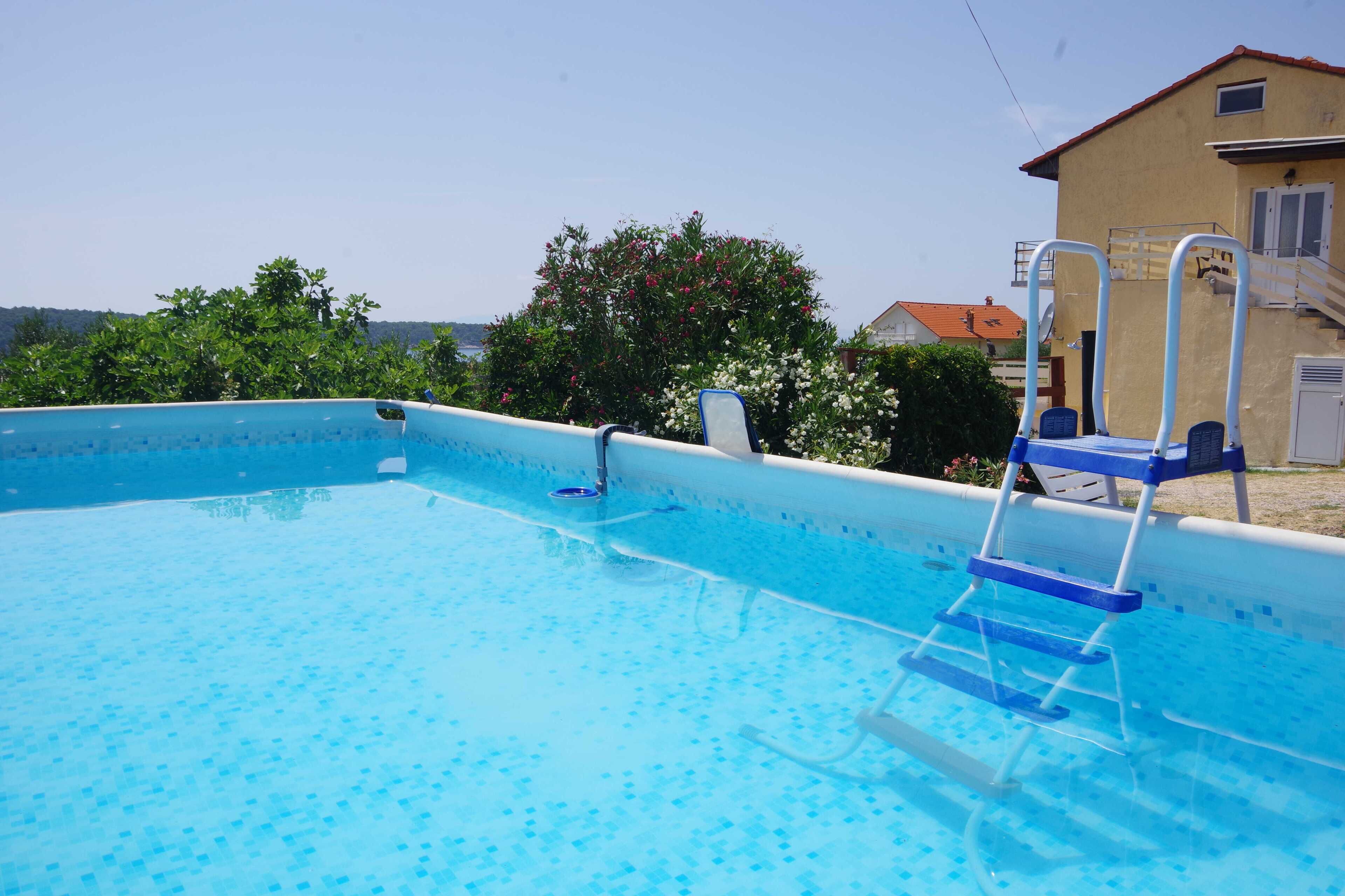 A clear blue swimming pool with a ladder, surrounded by greenery and a building with a red roof.