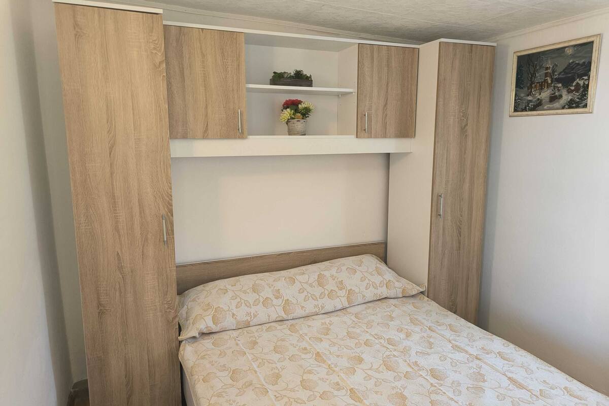 A bedroom with a bed, wooden wardrobe, and a shelf with plants.