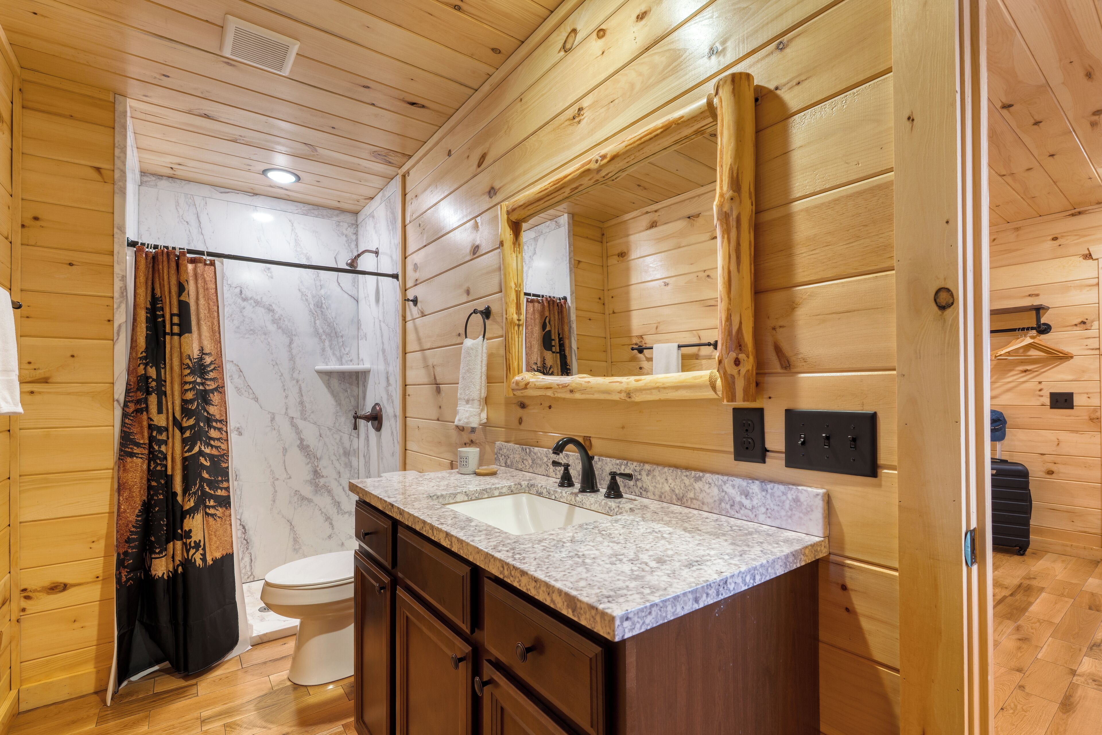 A wooden cabin bathroom with a marble sink, wooden cabinets, a large mirror, and a shower curtain with a forest design.