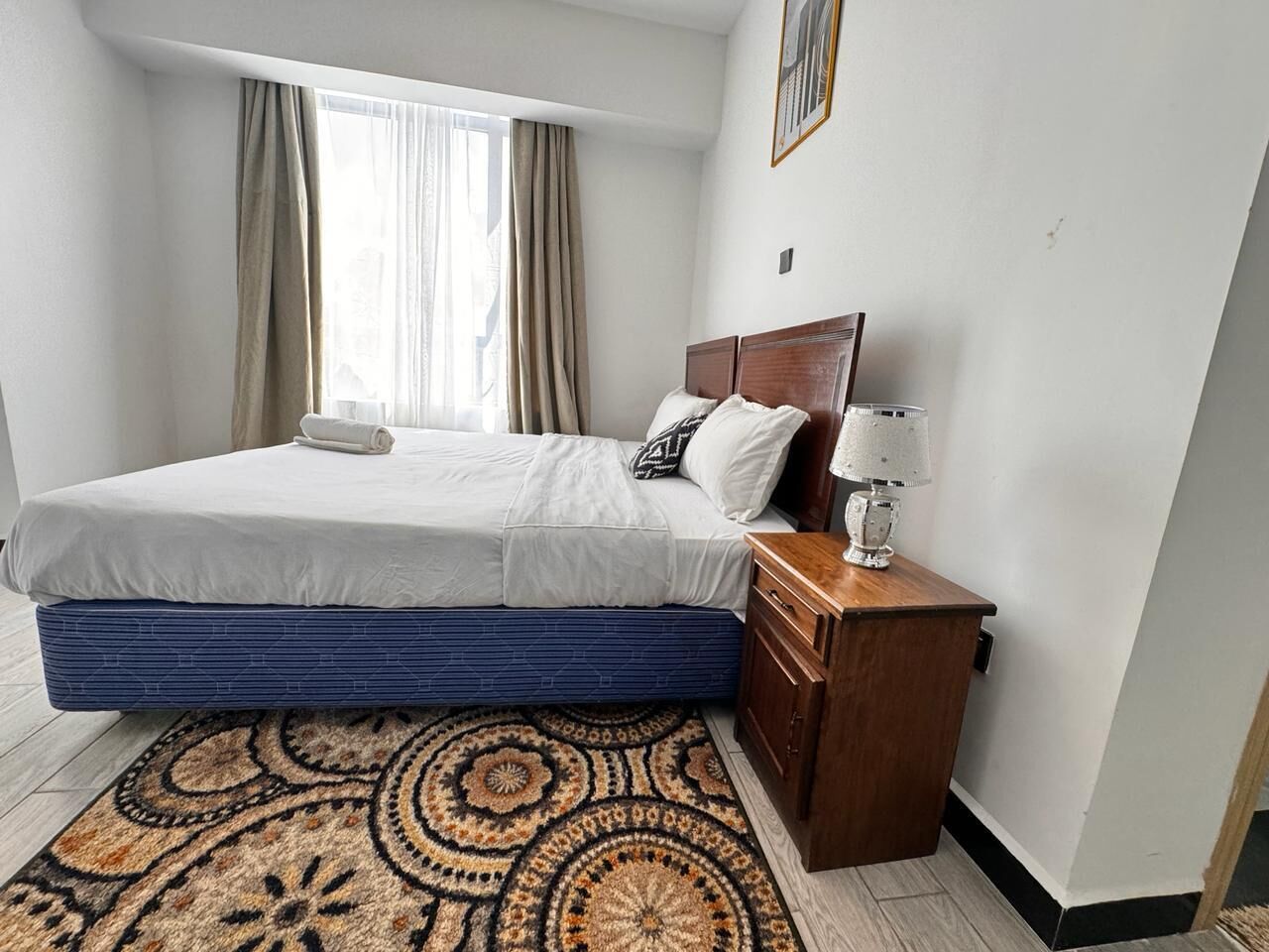 A hotel room with a bed, a nightstand, a lamp, a window with curtains, and a patterned rug.