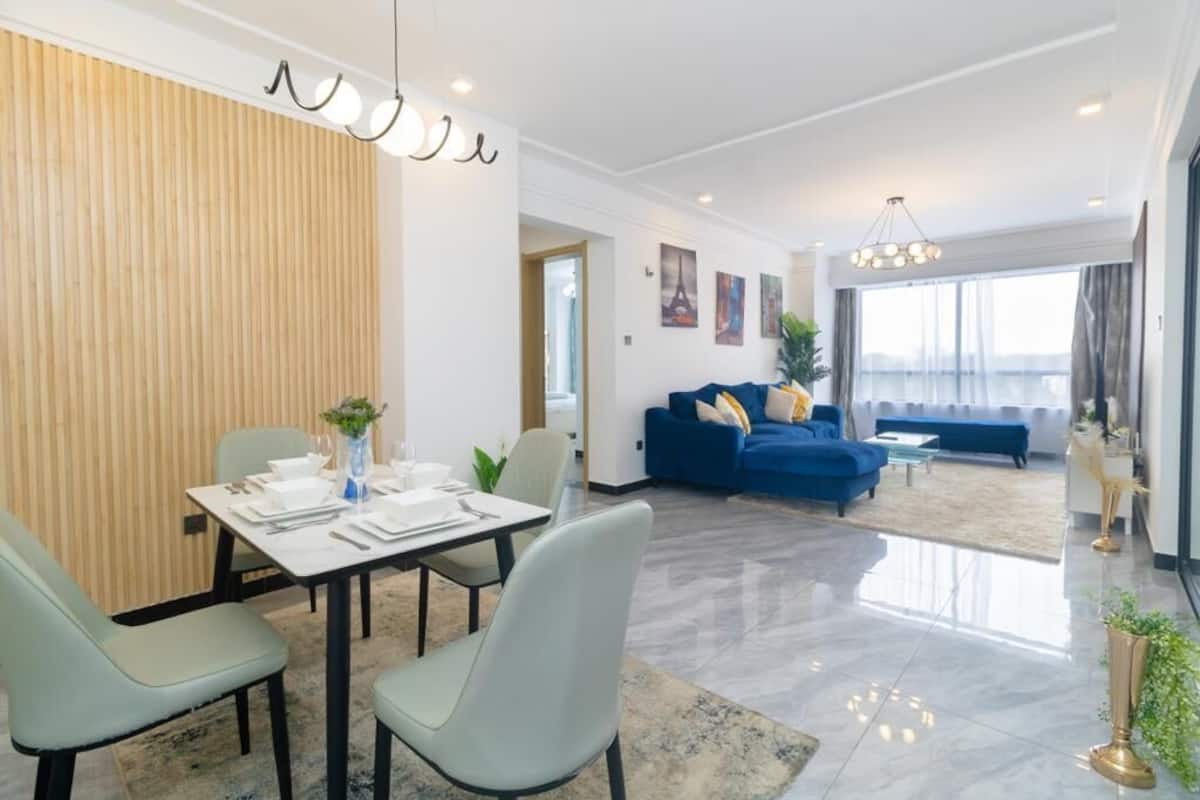 Una sala de estar moderna con un sofá azul, un comedor con una mesa preparada para cuatro personas y un gran ventanal con cortinas.