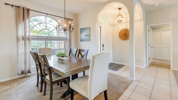 A dining area with a wooden table, chairs, and a chandelier. There is a large window with curtains, a door with a doormat, and a wall-mounted light fixture.