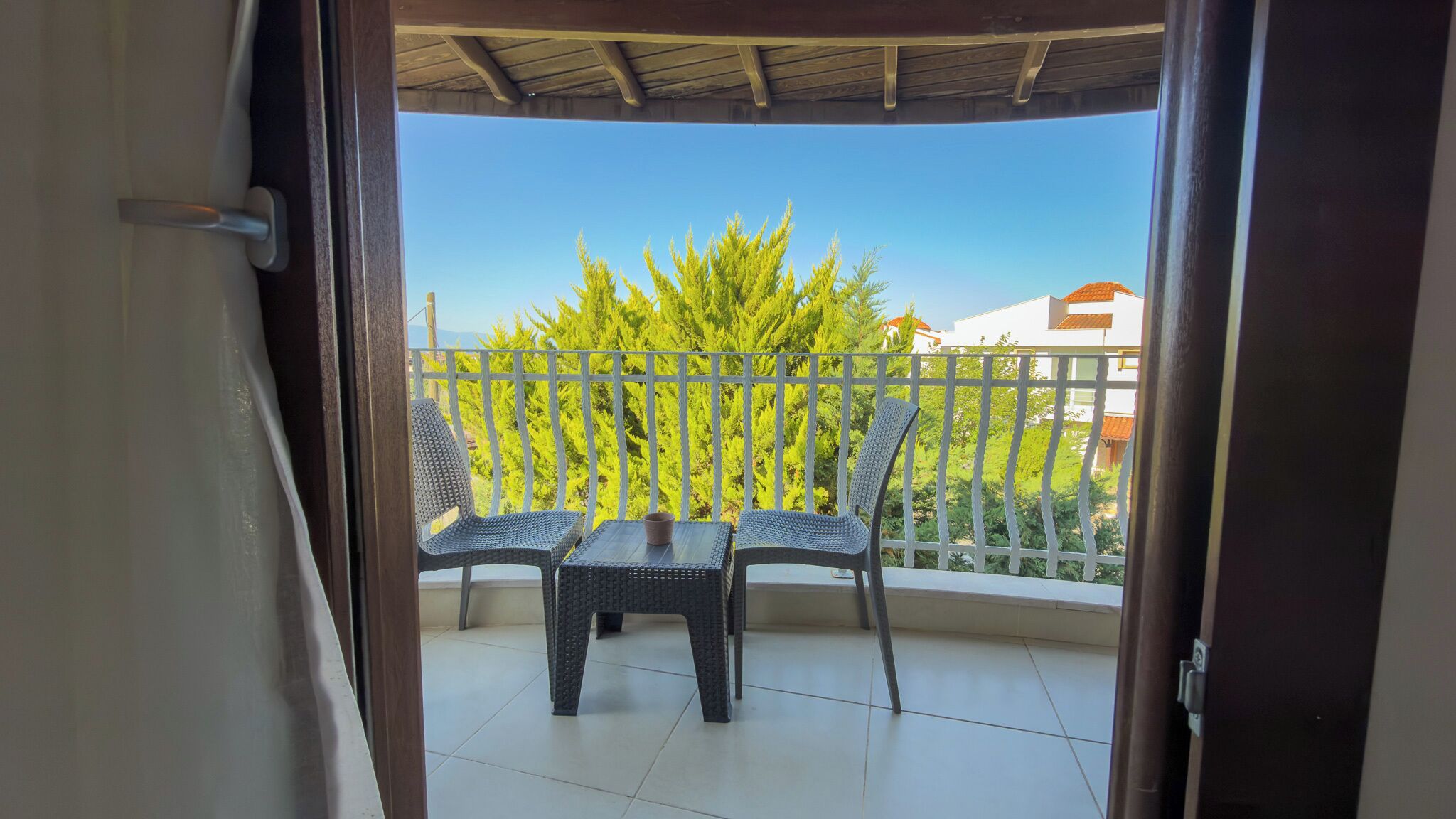 Un balcon ouvert avec deux fauteuils en osier et une petite table, une balustrade blanche, et une vue sur des arbres verdoyants et des bâtiments.