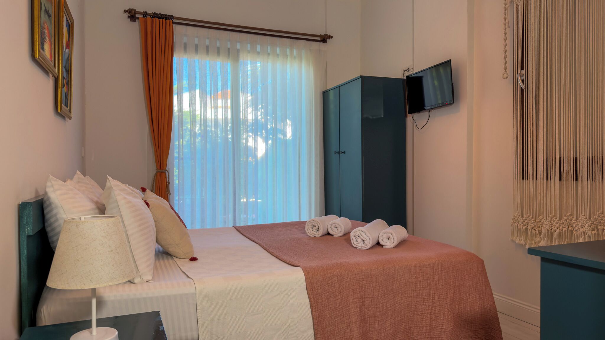 A bedroom with a bed, pillows, a bedside lamp, a TV, and a window with curtains.