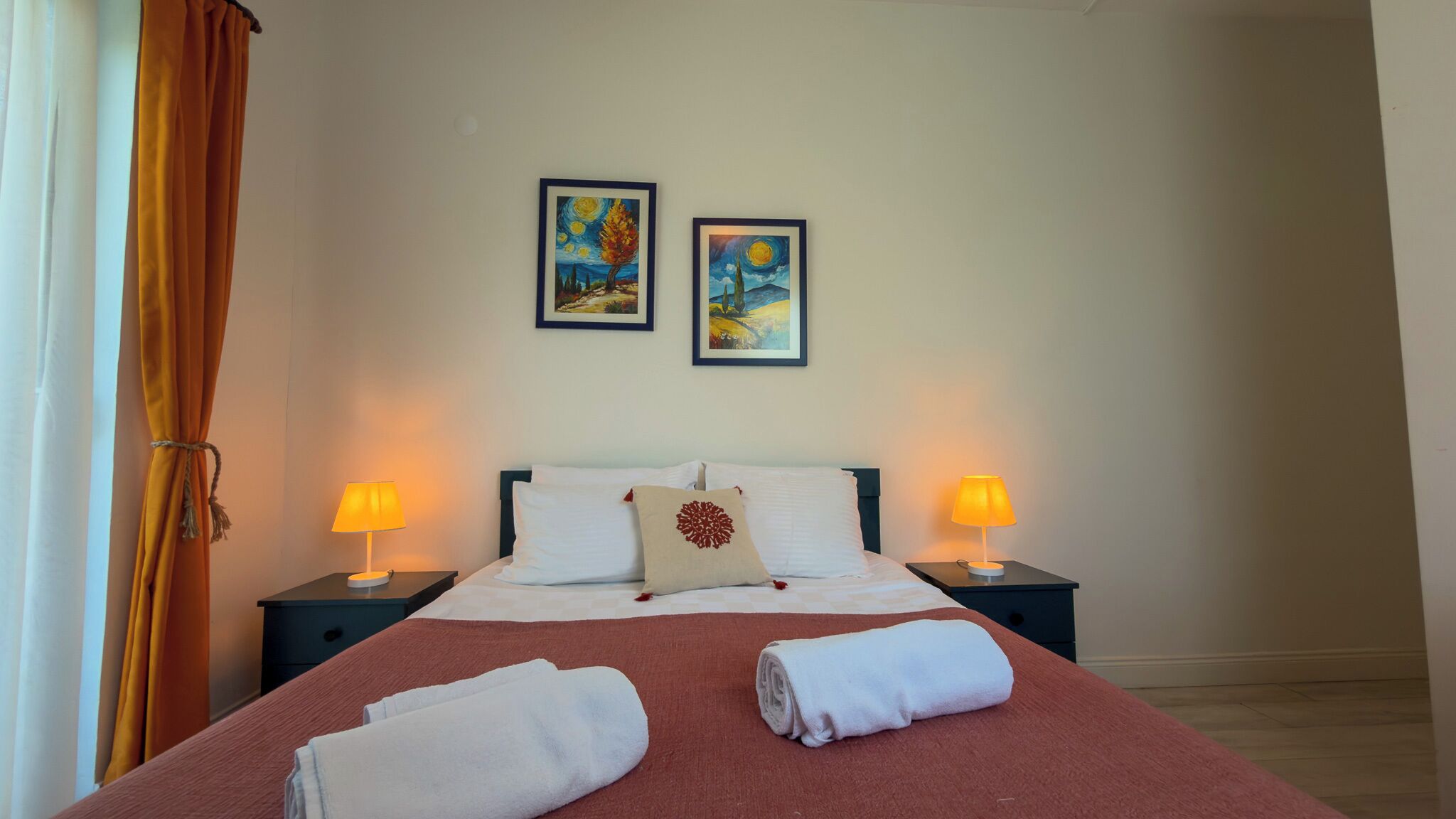 A hotel room with a bed, two bedside lamps, a window with curtains, and two framed paintings on the wall.