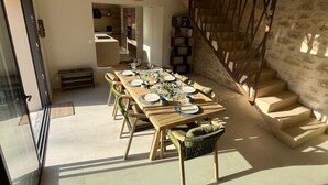 Un espace repas avec une longue table en bois, dressée pour six personnes, entourée de chaises, avec un escalier et un mur en pierre en arrière-plan.