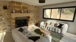 A living room with a stone fireplace, a sofa set, and a large window providing a view outside. - Villa Julorian near St Emilion (Sainte-Colombe)