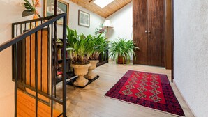 Una escalera de madera con barandal negro, plantas en macetas, un tapete rojo estampado y una puerta de madera.