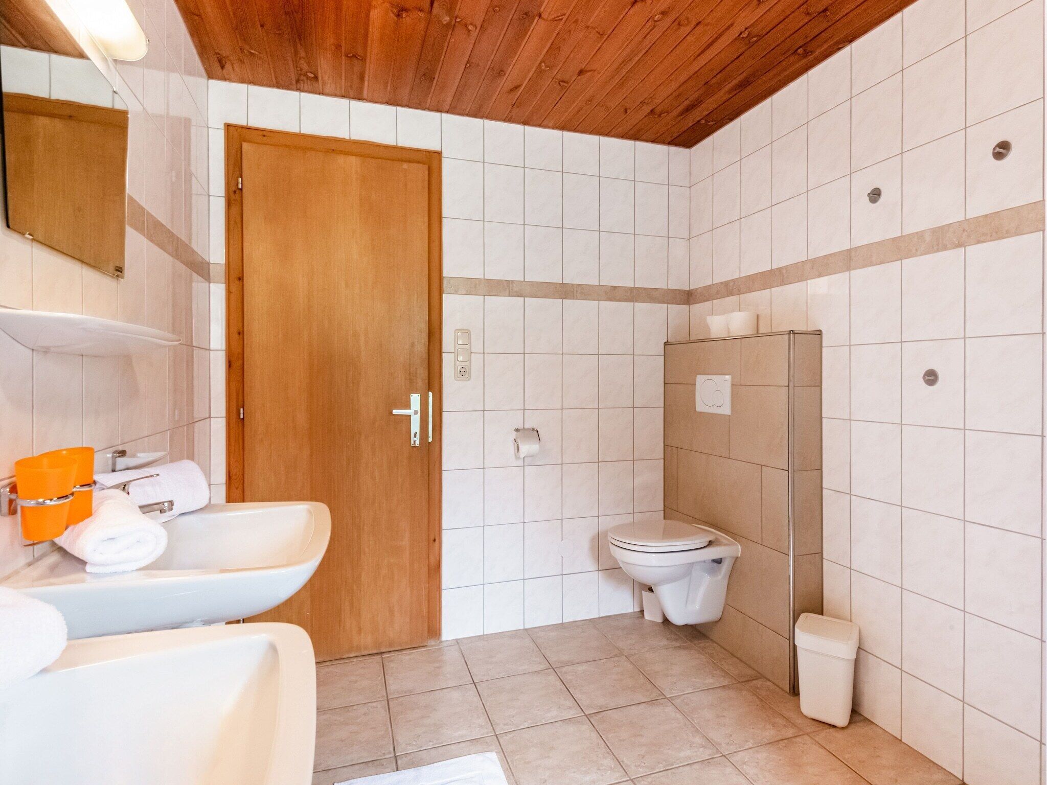 Un baño con puerta de madera, azulejos blancos, un lavamanos, un inodoro y una ducha.