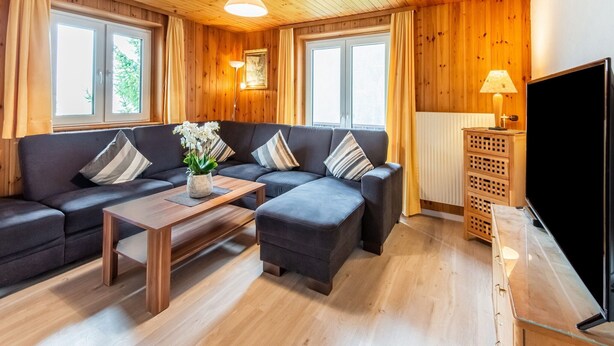 A cozy living room with a wooden ceiling, a dark grey sofa set, a wooden coffee table, and a large flat-screen TV.