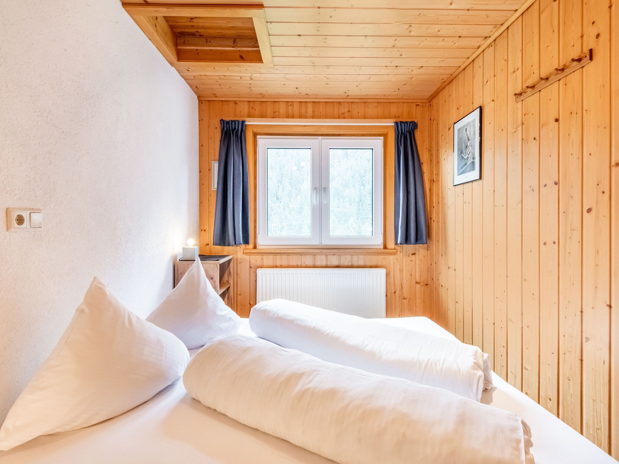 Ein Zimmer in einer Holzhütte mit zwei Betten, einem fenster mit Vorhängen, einem Heizkörper und einem gerahmten Bild an der Wand.