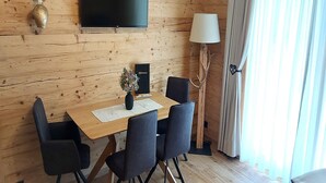 Een kamer met houten lambrisering, een houten tafel, vier stoelen, een flatscreen televisie, een lamp en een raam met gordijnen.