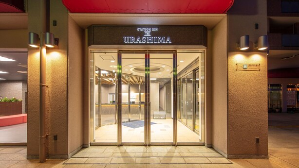 The entrance to Station Inn The Urashima, featuring a modern glass door entrance, a reception desk inside, and a red awning above. - STATION INN THE URASHIMA (Onomichi)