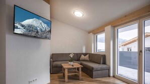 A modern living room with a wooden floor, a gray sectional sofa, a small wooden table with a vase, and a large window with a view of a snowy mountain. - Apartment Brixen near Ski Lift (Brixen im Thale)