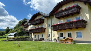 A hotel with multiple balconies adorned with red flowers, a well-maintained lawn, and a clear blue sky. - Apartment in Lungau near Ski Lift (St Michael im Lungau)