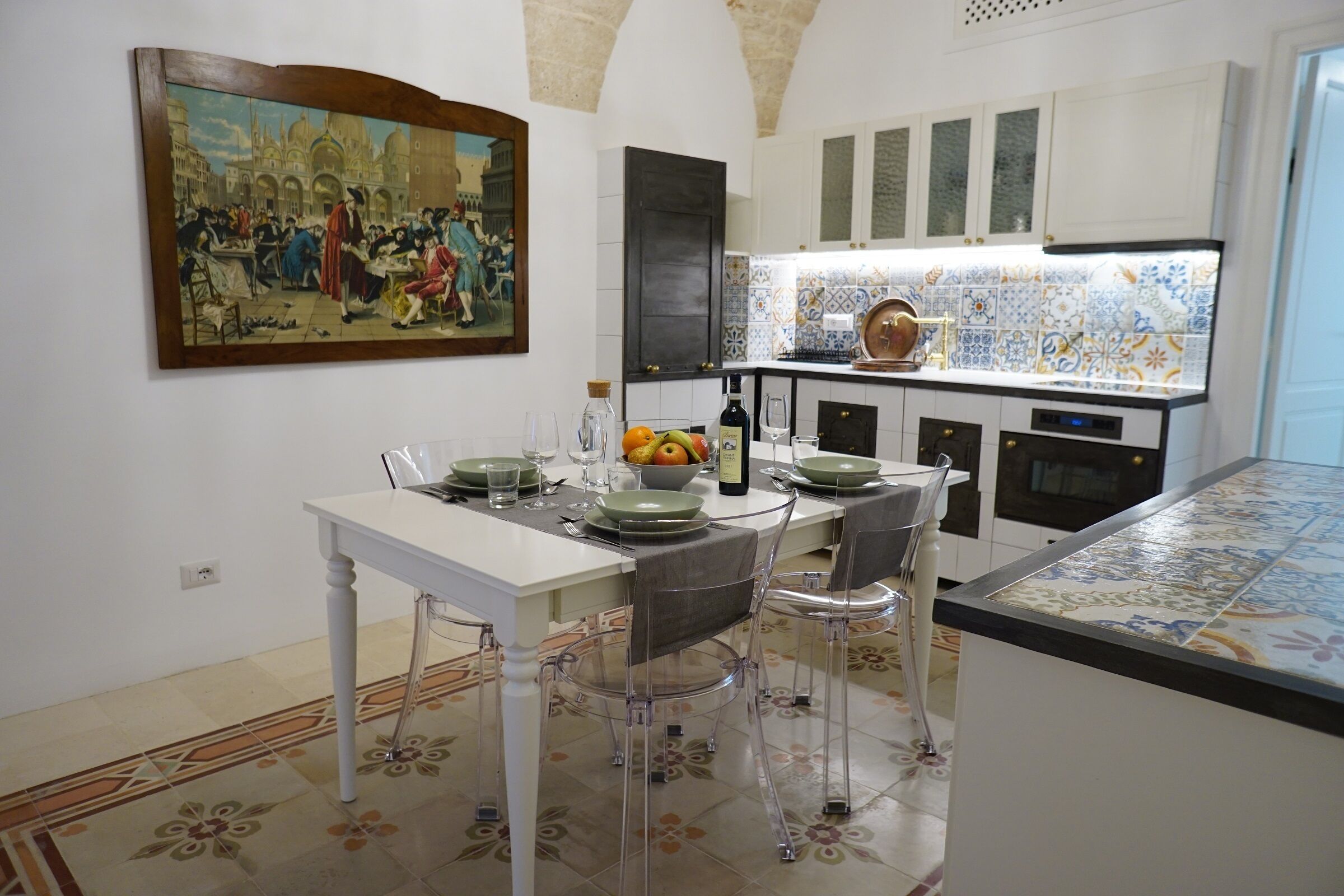 Un comedor con una mesa lista para cuatro personas, un cuadro en la pared y una cocina con azulejos estampados y armarios.