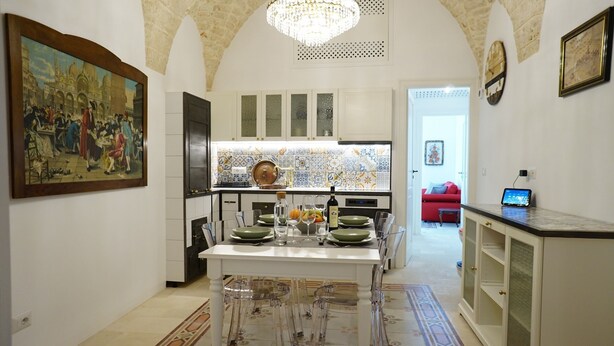 A dining area with a table set for four, clear chairs, a chandelier, a painting on the wall, and a kitchen in the background. - Heart of Puglia - Design Apartment in Puglia with 2 bedrooms (Putignano)