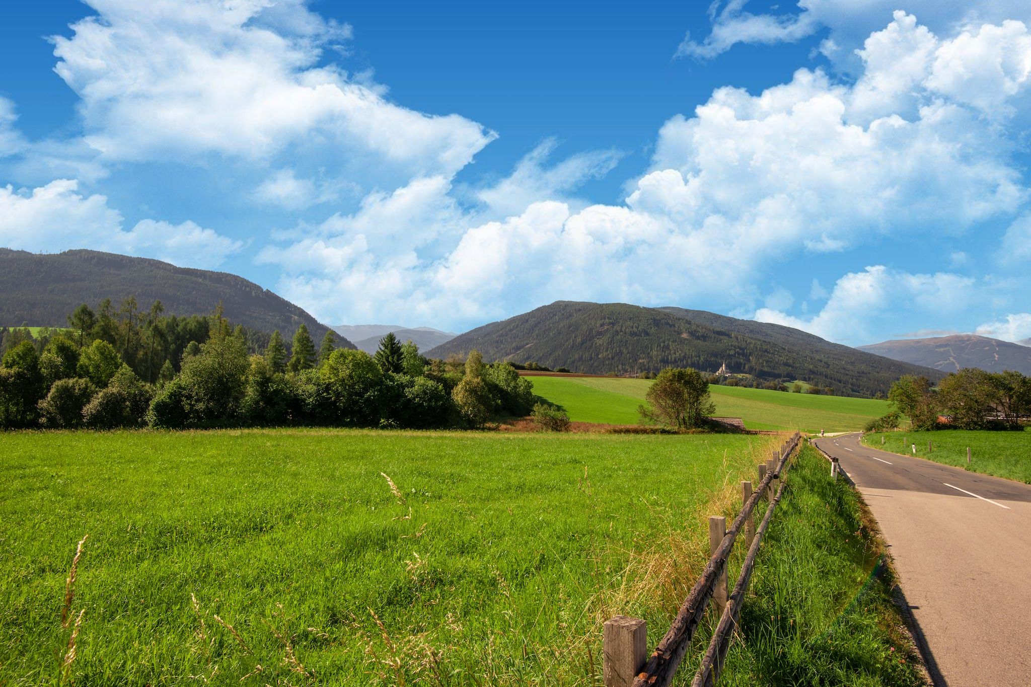 A rural road flanked by green fields and hills under a clear sky.