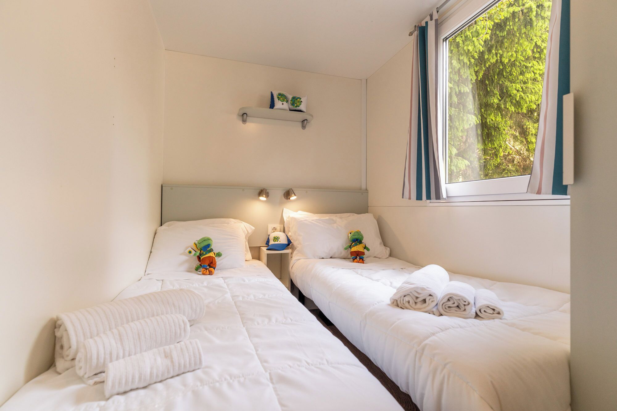 Habitación doble con dos camas, una mesita de noche con lámpara, un estante con objetos decorativos y una ventana con vista a la vegetación.