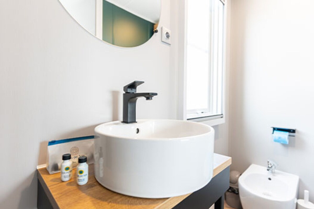 Un bagno moderno con un lavandino bianco grande, un lavandino bianco più piccolo e uno specchio sopra il lavandino più grande. Ci sono due bottiglie sul ripiano.