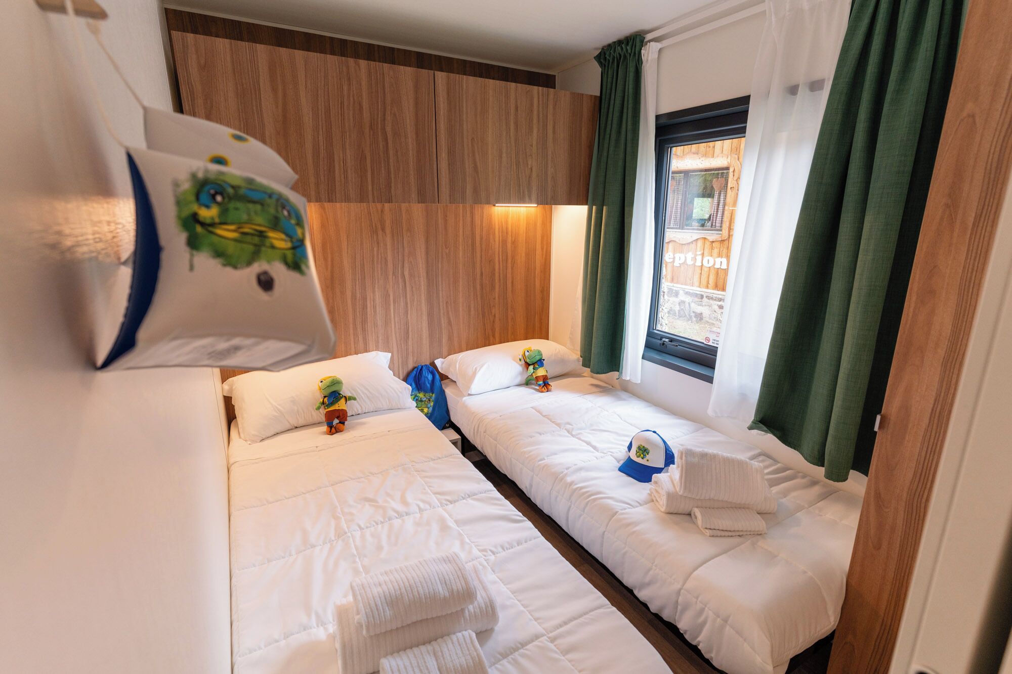 Hotellihuone, jossa on kaksi erillistä sänkyä, puinen pääty, ikkuna verhoilla ja pieni pöytä, jonka päällä on lelu.