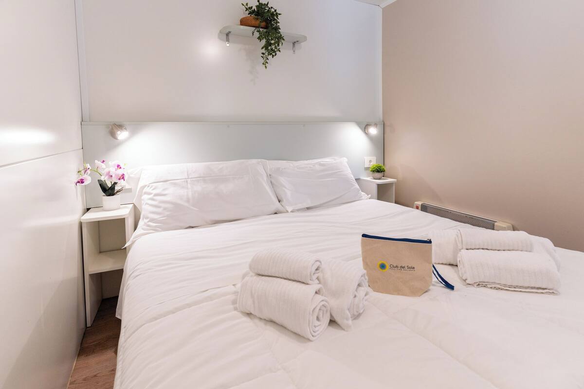 A neatly made bed with white linens, a folded towel, and a bag labeled 'Club del Sole' on the bedside table.