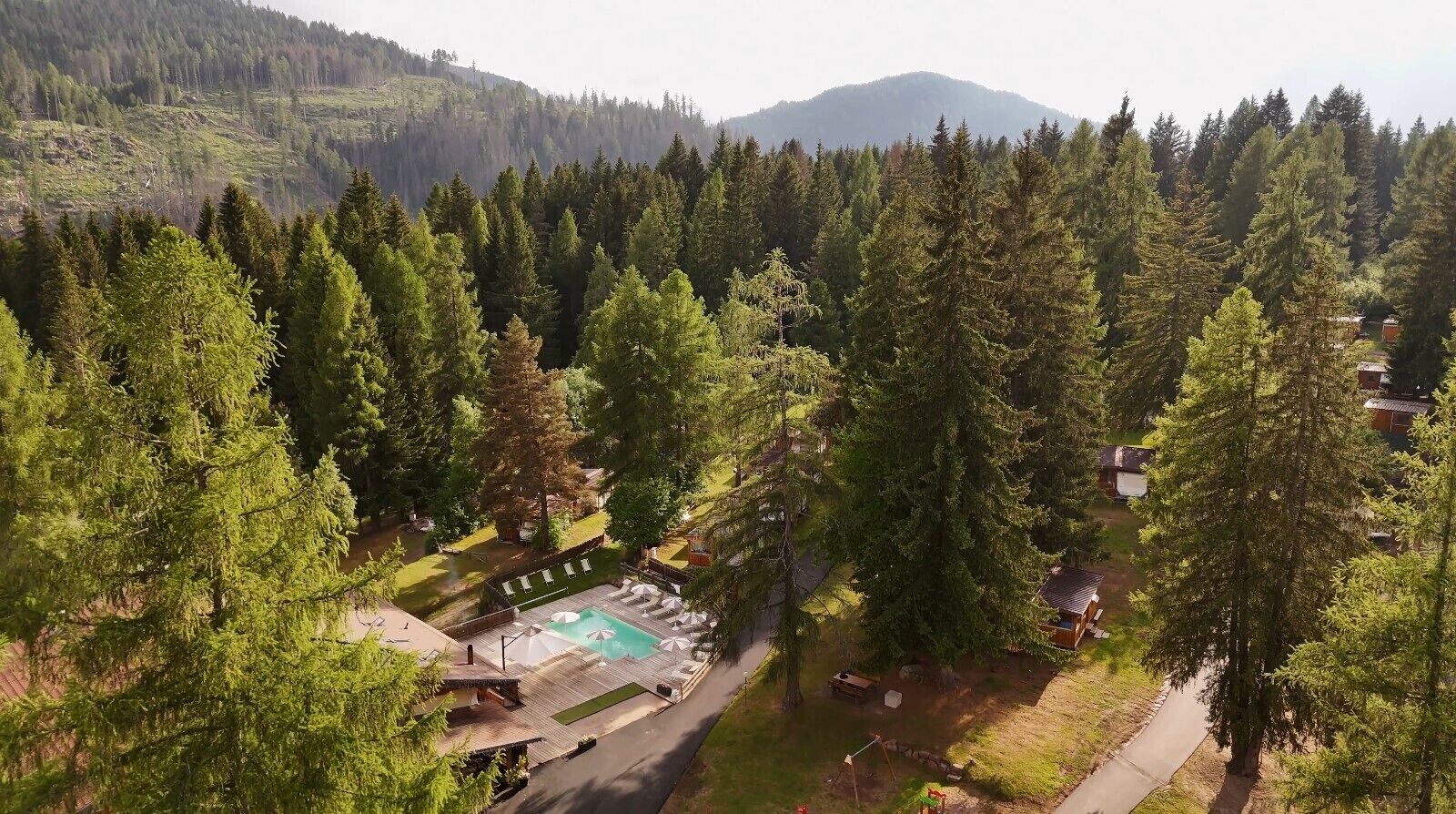 An aerial view of a resort with a swimming pool surrounded by trees, a small building, and a mountain in the background.