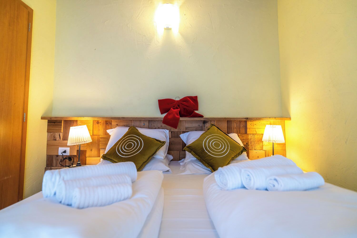 Une chambre d’hôtel avec deux lits, une tête de lit en bois, deux lampes de chevet et un nœud rouge décoratif.