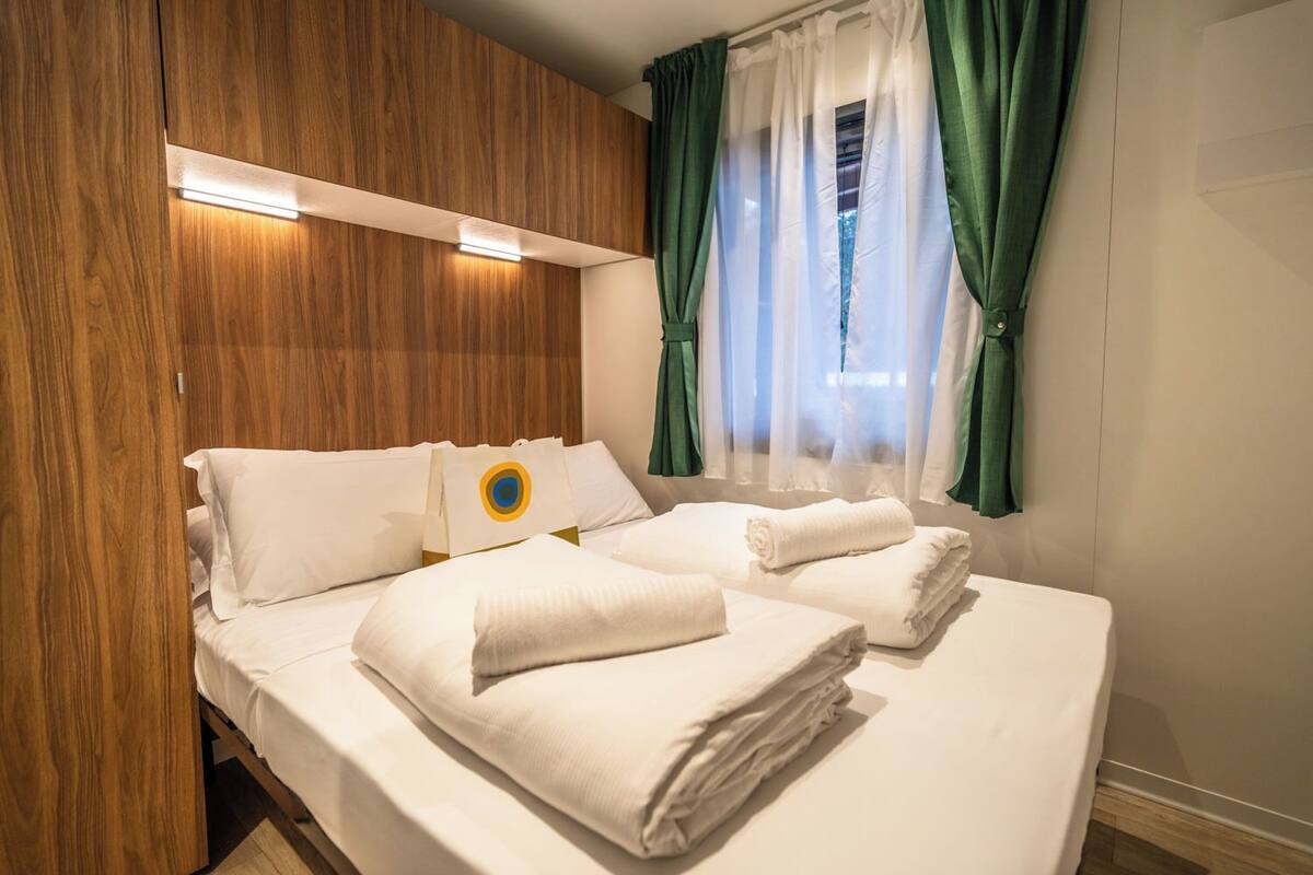 A hotel room with a wooden headboard, a bed with white linens, a small table with a towel, and a window with green curtains.